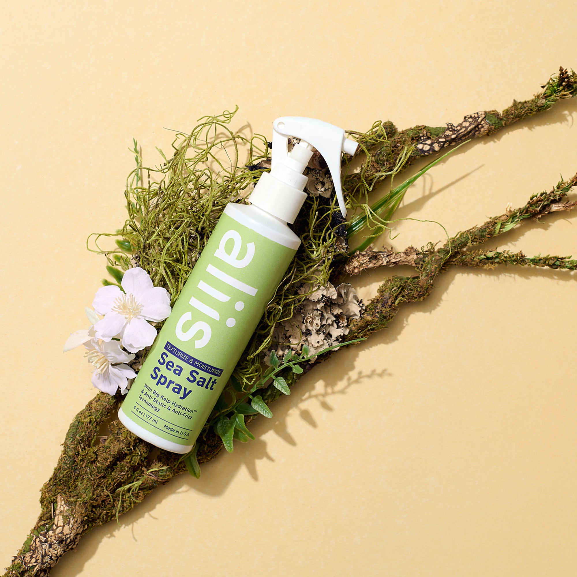 Green spray bottle labeled Aliis on a natural background with plants and flowers.